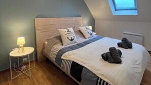 A bedroom with a bed, a bedside lamp, a small table, a window, and a radiator. - Ty Manoa - Pretty house 50m from the sea (Landéda)