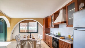 A kitchen with a dining table set for four, wooden cabinets, a refrigerator, and a window overlooking a backyard. - Sirena Apartment in Luxury Villa with Shared Pool (Scala di Furno)