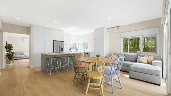 A modern kitchen with a central island, a dining area with mismatched chairs, and a grey sofa in a bright living room with large windows.