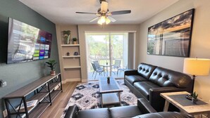 A living room with a leather sofa, a coffee table, a TV mounted on the wall, a ceiling fan, and a sliding glass door leading to an outdoor area. - 12-204 Legacy Dunes, Free WiFi, Heated Pool, Close to Disney (Kissimmee)