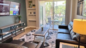 A living room with a TV, sofa, coffee table, and a balcony with chairs. - 12-204 Legacy Dunes, Free WiFi, Heated Pool, Close to Disney (Kissimmee)