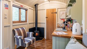 A cozy cabin interior with a wood-burning stove, a striped chair, a kitchen area with a coffee maker, and a window with a view of trees.