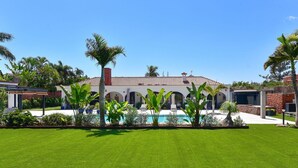 Una casa con piscina, rodeada de césped cuidado y palmeras.