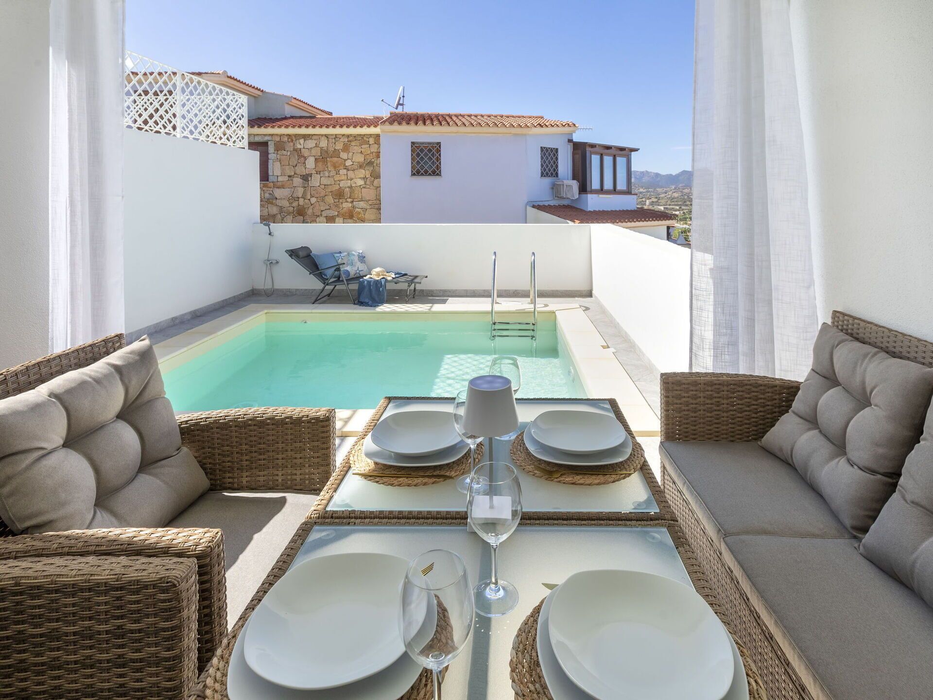 Un espace repas aménagé au bord de la piscine, avec des meubles en osier, une table dressée pour deux personnes et une vue sur un bâtiment doté d’une façade en pierre et d’un toit en tuiles.