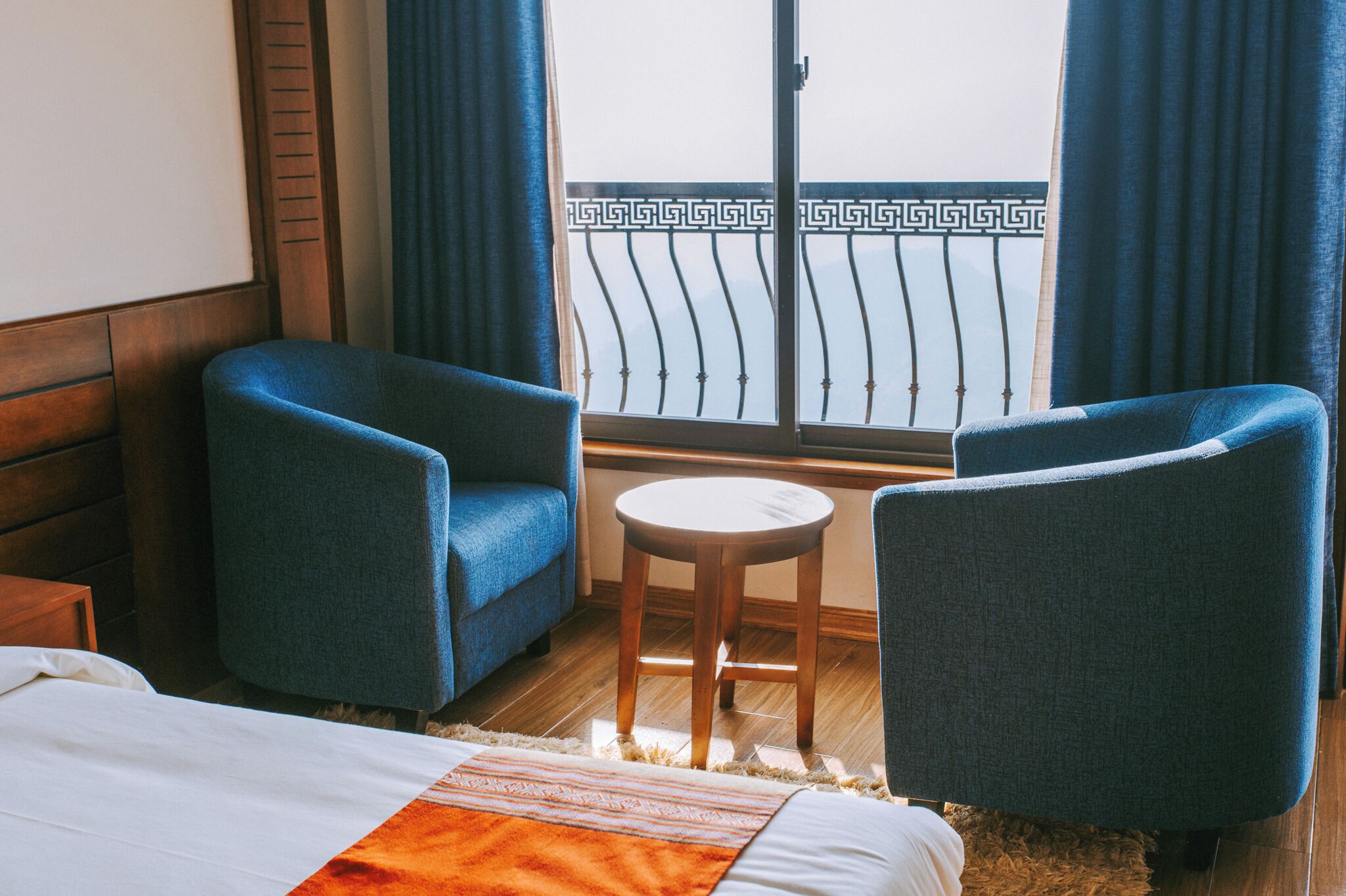 A hotel room with a bed, two blue armchairs, a small wooden table, and a view of a balcony with a decorative railing.