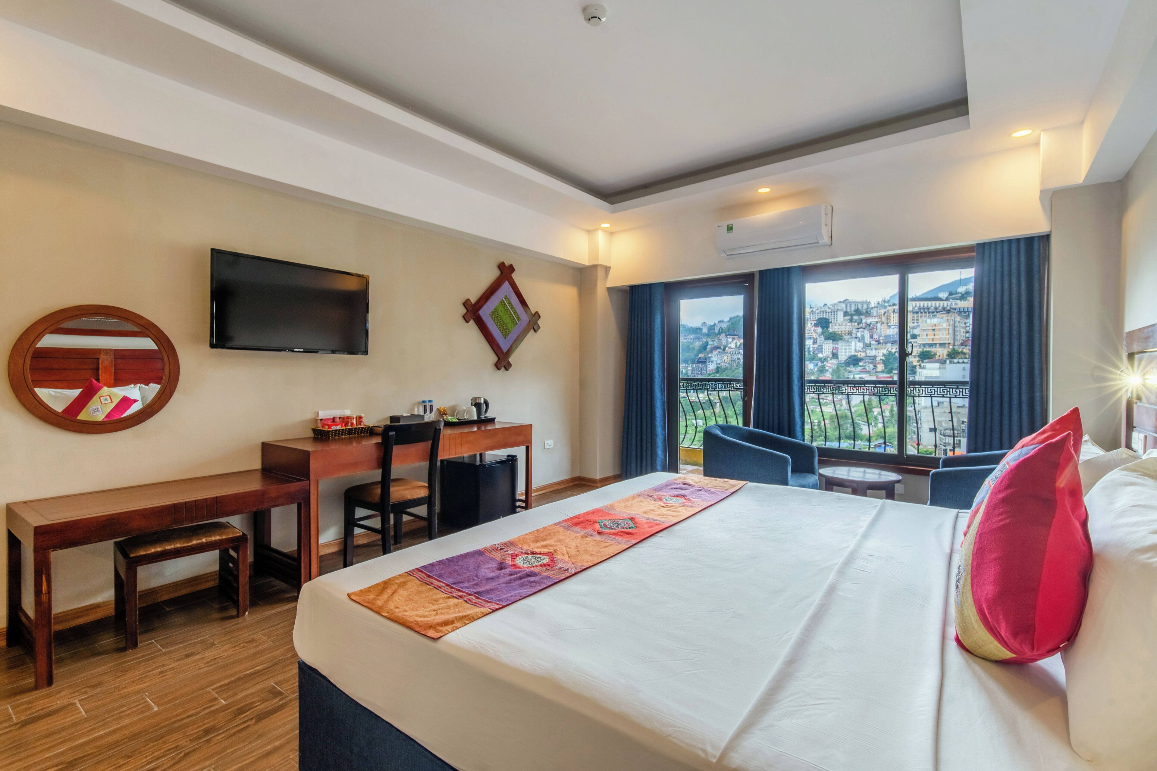 A hotel room with a bed, desk, chair, TV, and a view of the city.