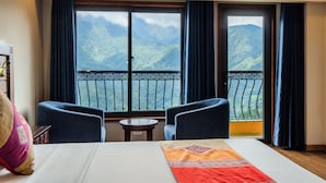 A hotel room with a bed, two armchairs, a small table, and a balcony with a mountain view. - Sapa Charm Hotel (Sa Pa)