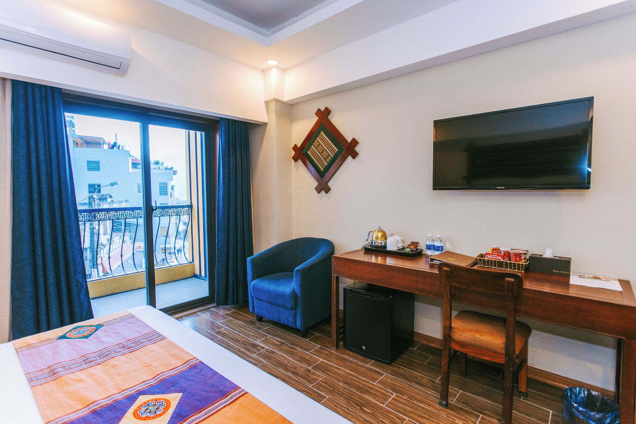 A hotel room with a bed, a desk, a chair, a TV, a small fridge, and a window with a view of buildings.