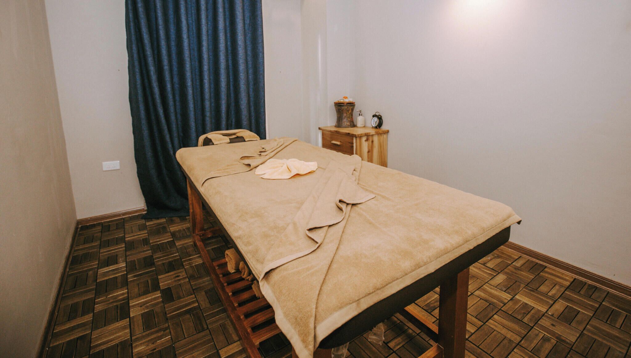 A room with a wooden bench, a table with a beige cloth, a small wooden side table with a vase and a clock, and a blue curtain.
