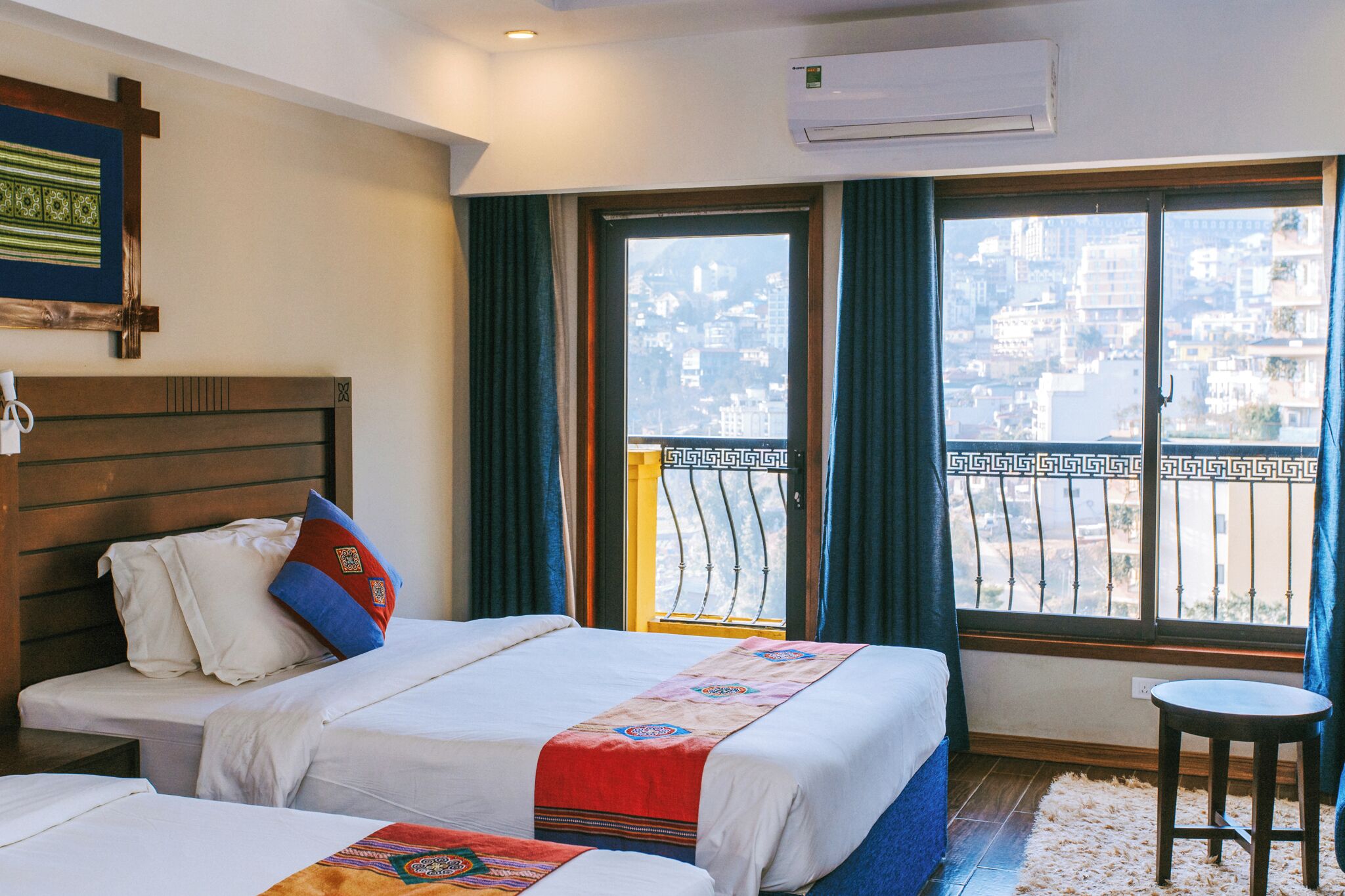 A hotel room with a bed, a small table, a balcony with a view of the city, and an air conditioning unit.