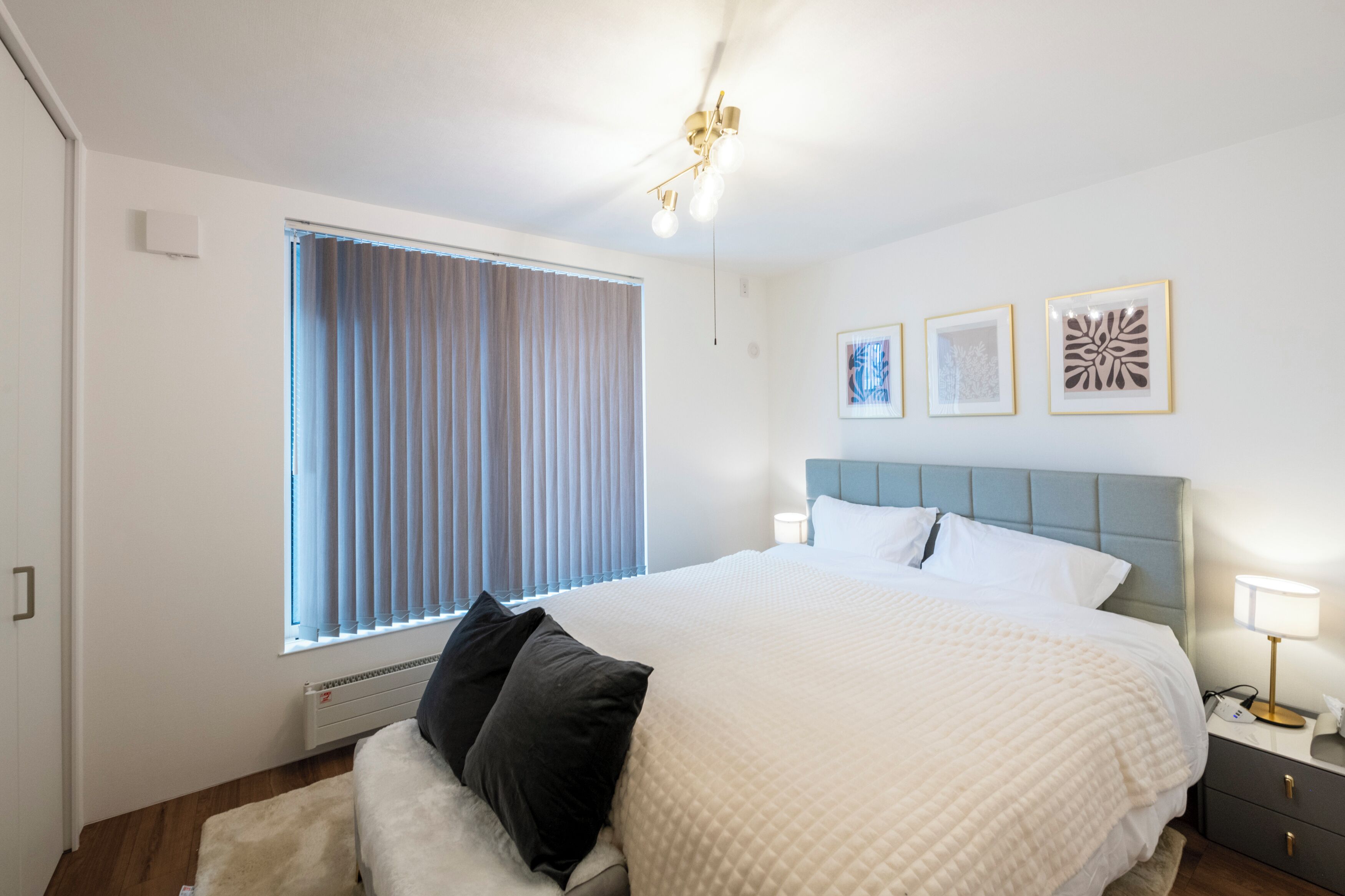 A bedroom with a large bed, a bedside table with a lamp, a window with blinds, and three framed pictures on the wall.
