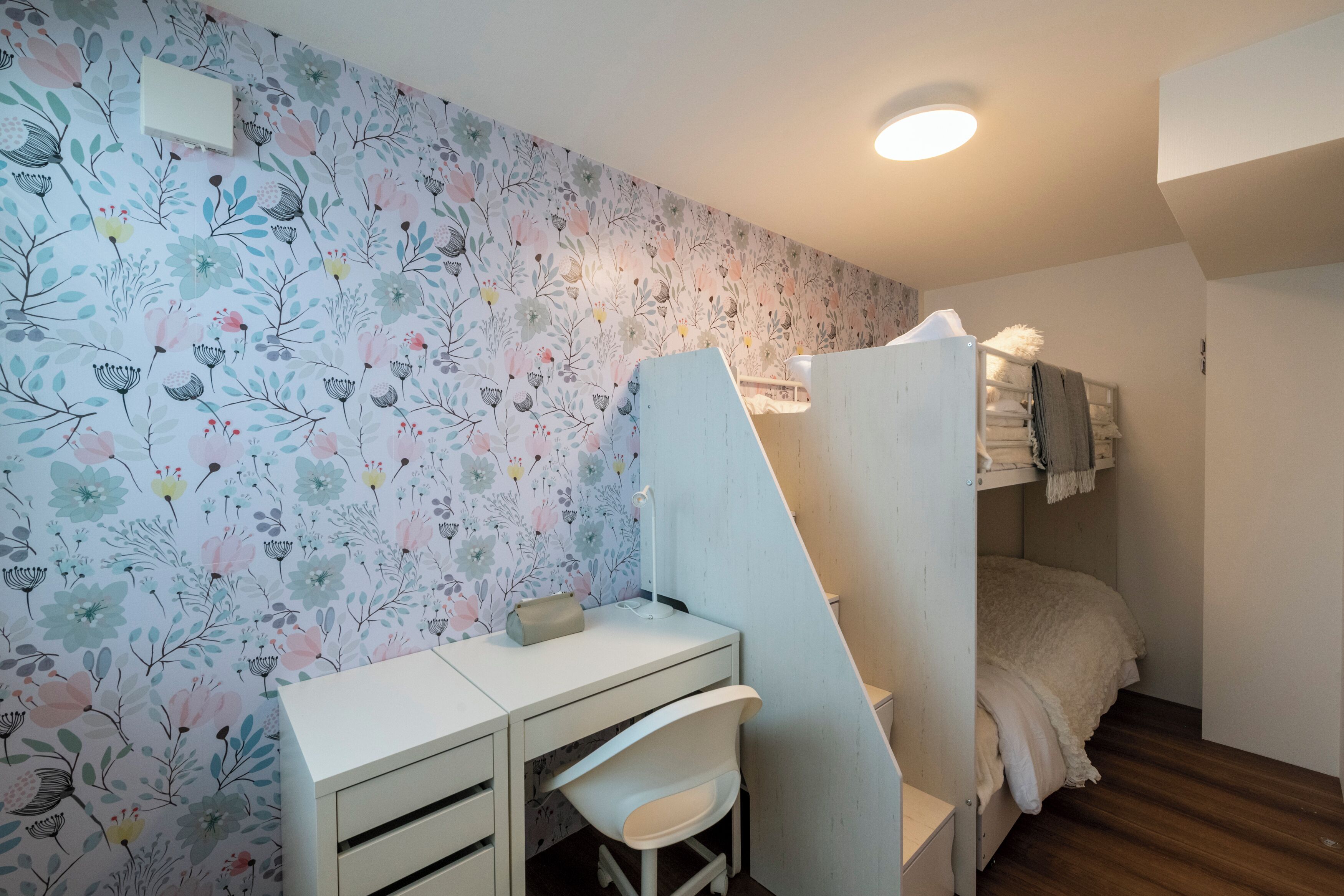 Un dormitorio con papel tapiz floral, un escritorio con una silla y una cama con una manta blanca.