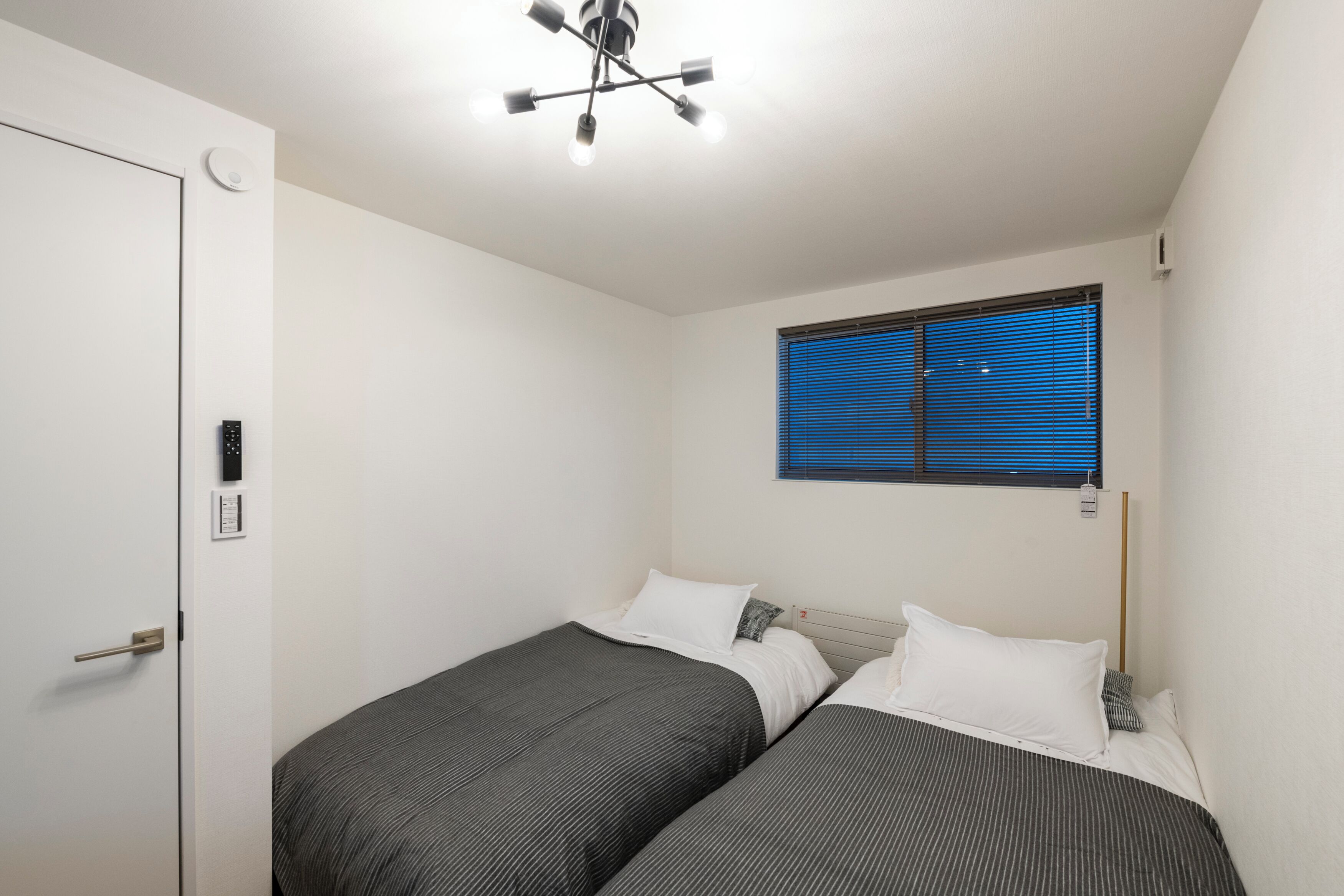 A small, clean room with two single beds, a white door, a ceiling light fixture, and a window with blinds.