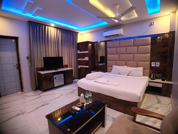 A modern hotel room with a large bed, a TV, a minibar, a desk with a remote, and a ceiling fan. - Samudra Bilas Resort (Kanthi)