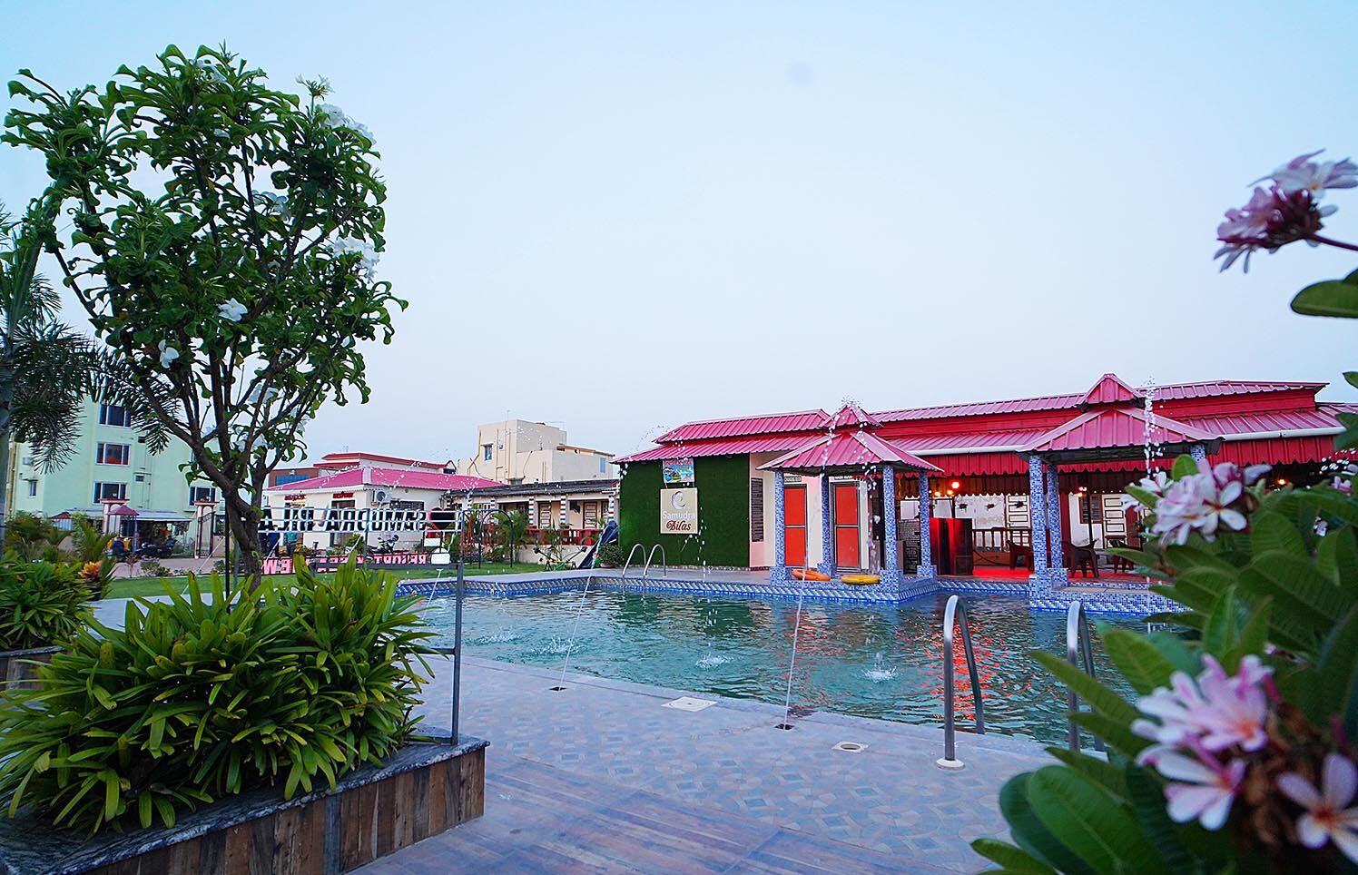 A swimming pool area with a tiled deck, surrounded by lush greenery and a building with red roofs.