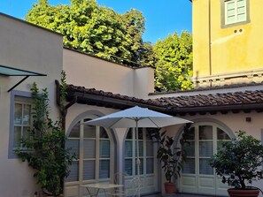 A courtyard with a table and chairs under a white umbrella, surrounded by potted plants and a building with arched windows. - Inside Lucca  Walls, Historic Home,  private Garden, Terrace (Lucca)