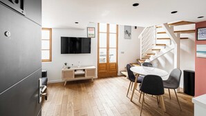 Un espace de vie moderne doté d’un plancher en bois, d’un coin repas avec une table ronde et des chaises, d’un téléviseur fixé au mur et d’un escalier avec des marches en bois.