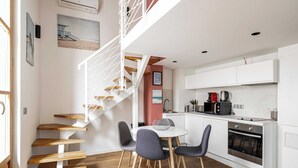 Une cuisine moderne avec des armoires blanches, un four encastré et une machine à café. On y trouve un coin repas avec une table ronde et quatre chaises. Un escalier blanc avec des marches en bois mène à l’étage supérieur.