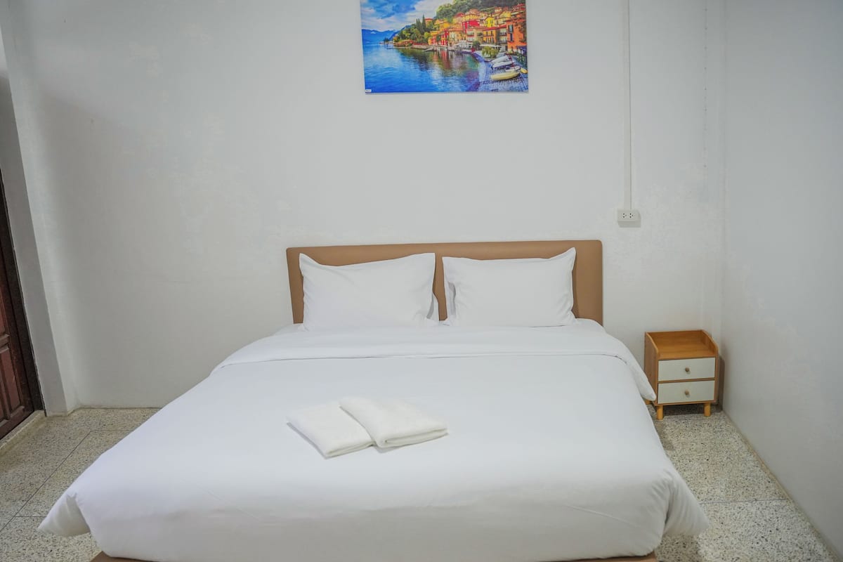 A neatly made bed with white linens and two folded towels, a wooden nightstand with a drawer, and a painting of a colorful coastal scene on the wall.