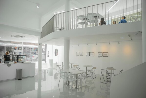 A spacious, modern interior with a central seating area, a balcony with tables and chairs, and a counter with a person behind it. - Scenario Cafe and Hostel (Kanchanaburi)