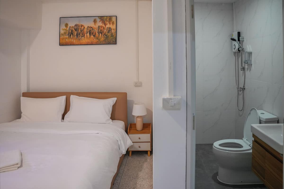 A hotel room with a bed, a nightstand, a lamp, a painting of elephants, and a bathroom with a toilet and shower.