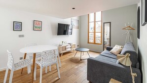 Un salon moderne avec un canapé gris, des chaises blanches, une table de salle à manger ronde et un téléviseur fixé au mur.