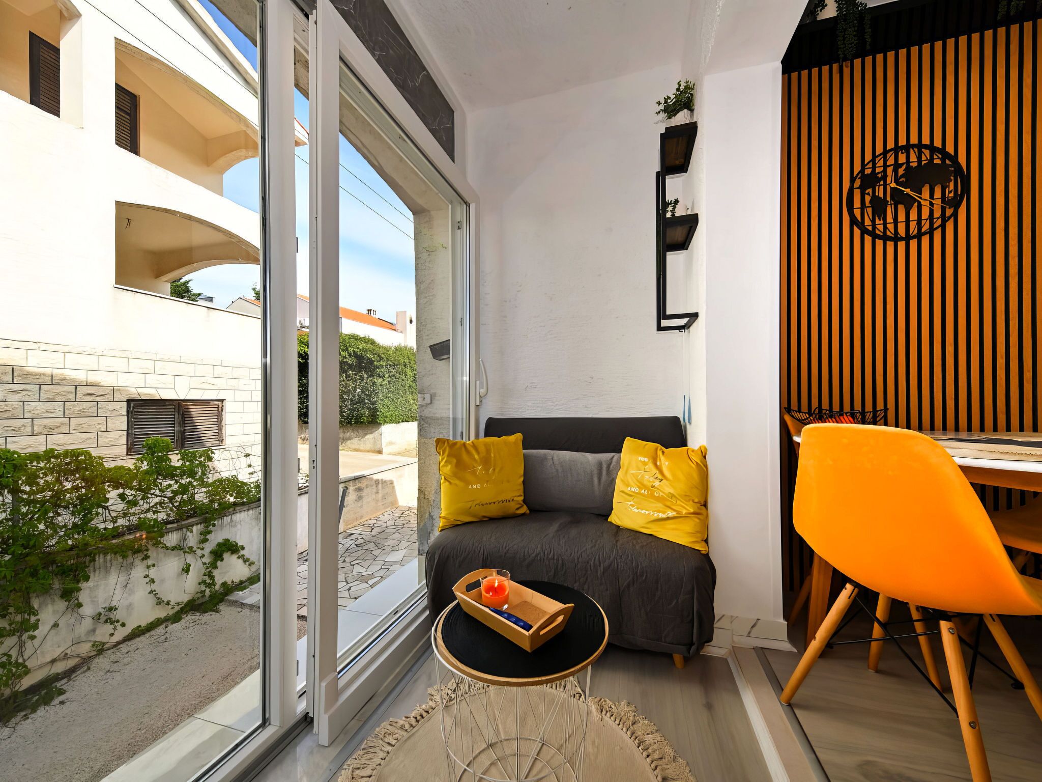 A modern living room with a sofa, a dining table, and a chair. There is a window with a view of a building and greenery outside.