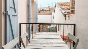 Ein schmaler Holzbalkon mit Metallgeländer, der zu einem Innenhof mit weißem Zaun und einem Gebäude mit rotem Dach führt.