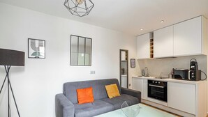 Ein modernes Wohnzimmer mit einem grauen Sofa, zwei Kissen, einem Glastisch und einer Küchenzeile mit weißen Schränken, einer Mikrowelle und einem Backofen.