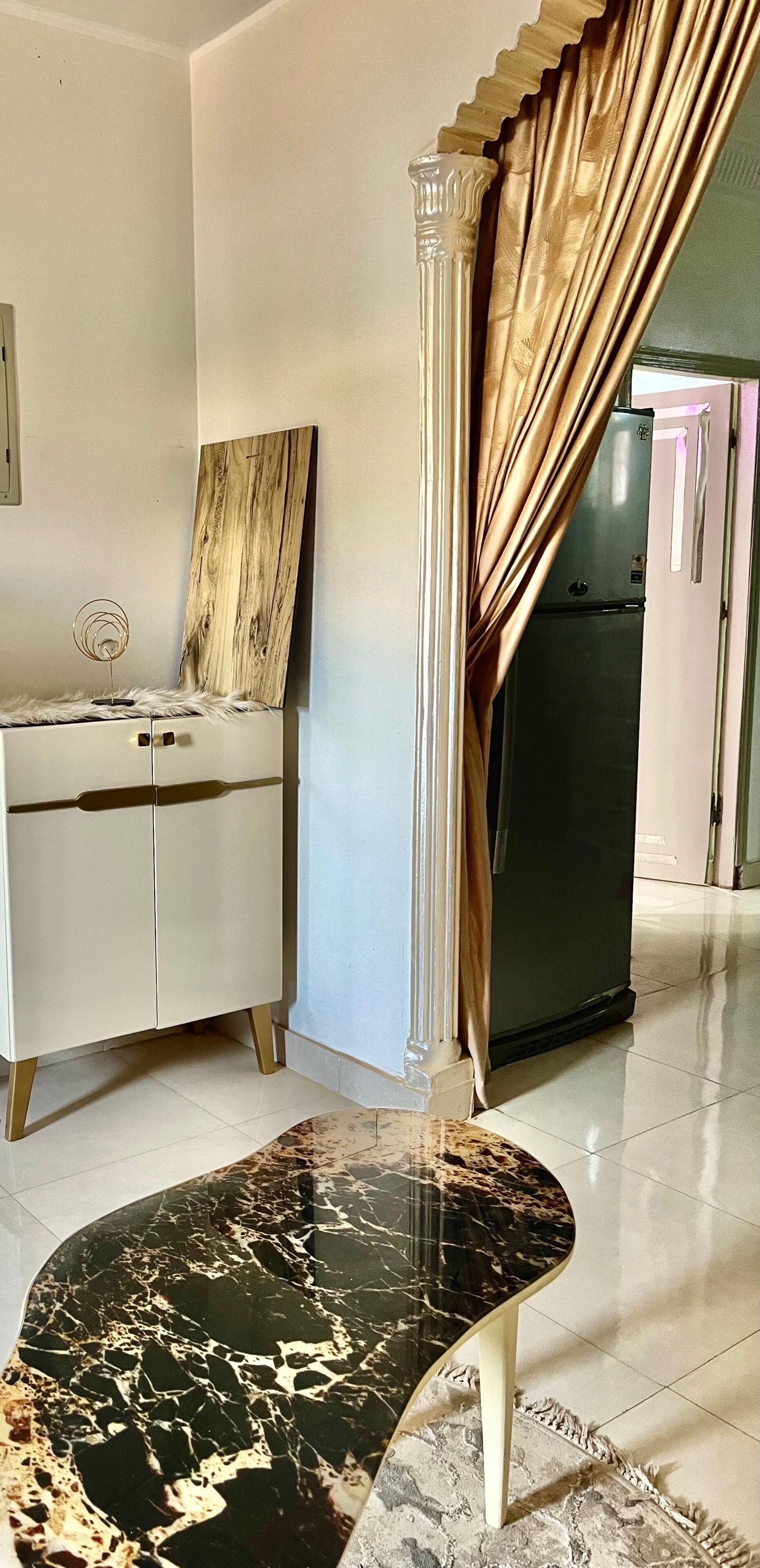 Una habitación con una mesa de mármol, un armario blanco, una obra de arte de madera, una columna y un refrigerador.