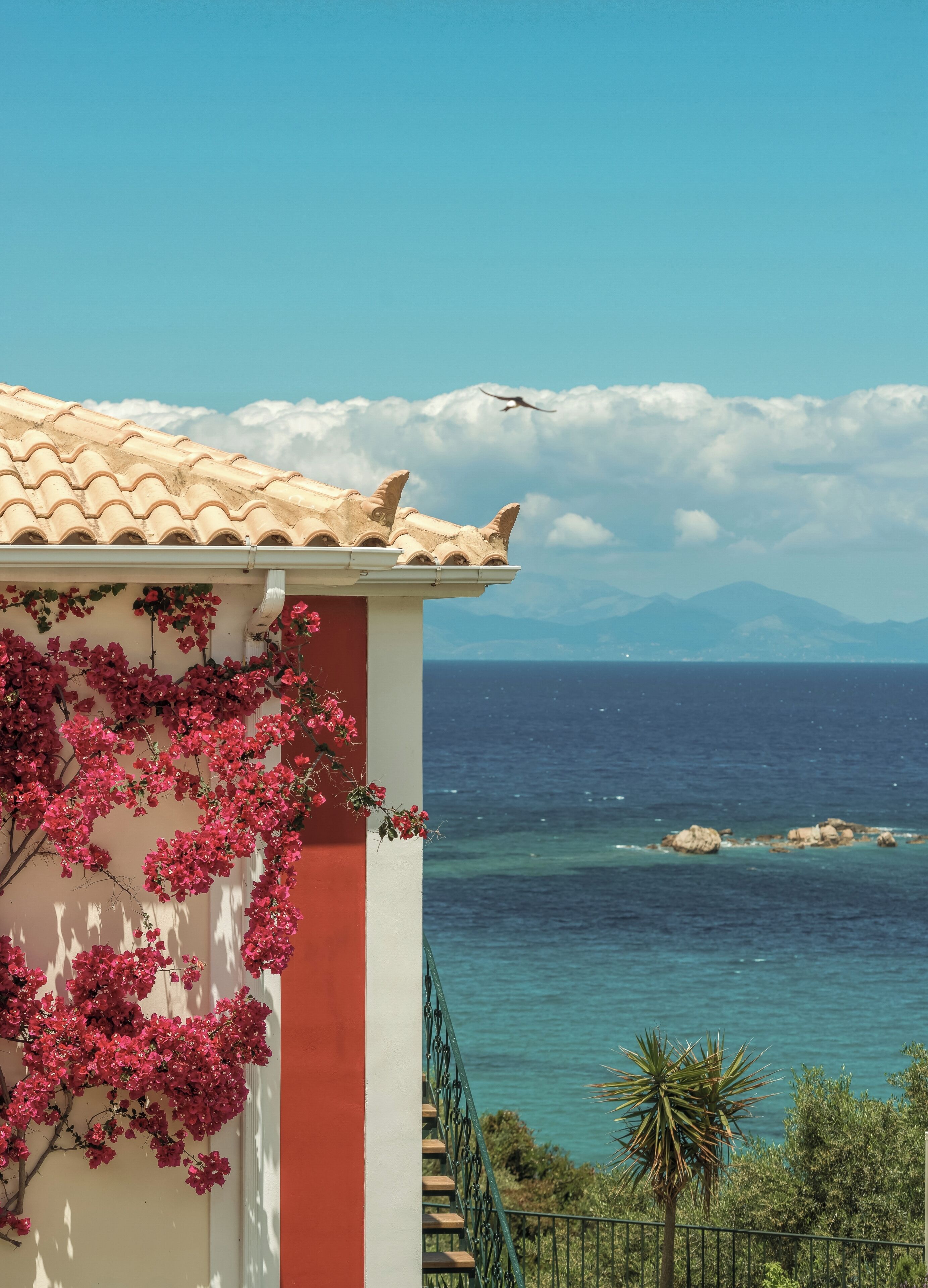 A coastal building with a tiled roof, pink flowering vines, and a view of the sea and mountains in the background.