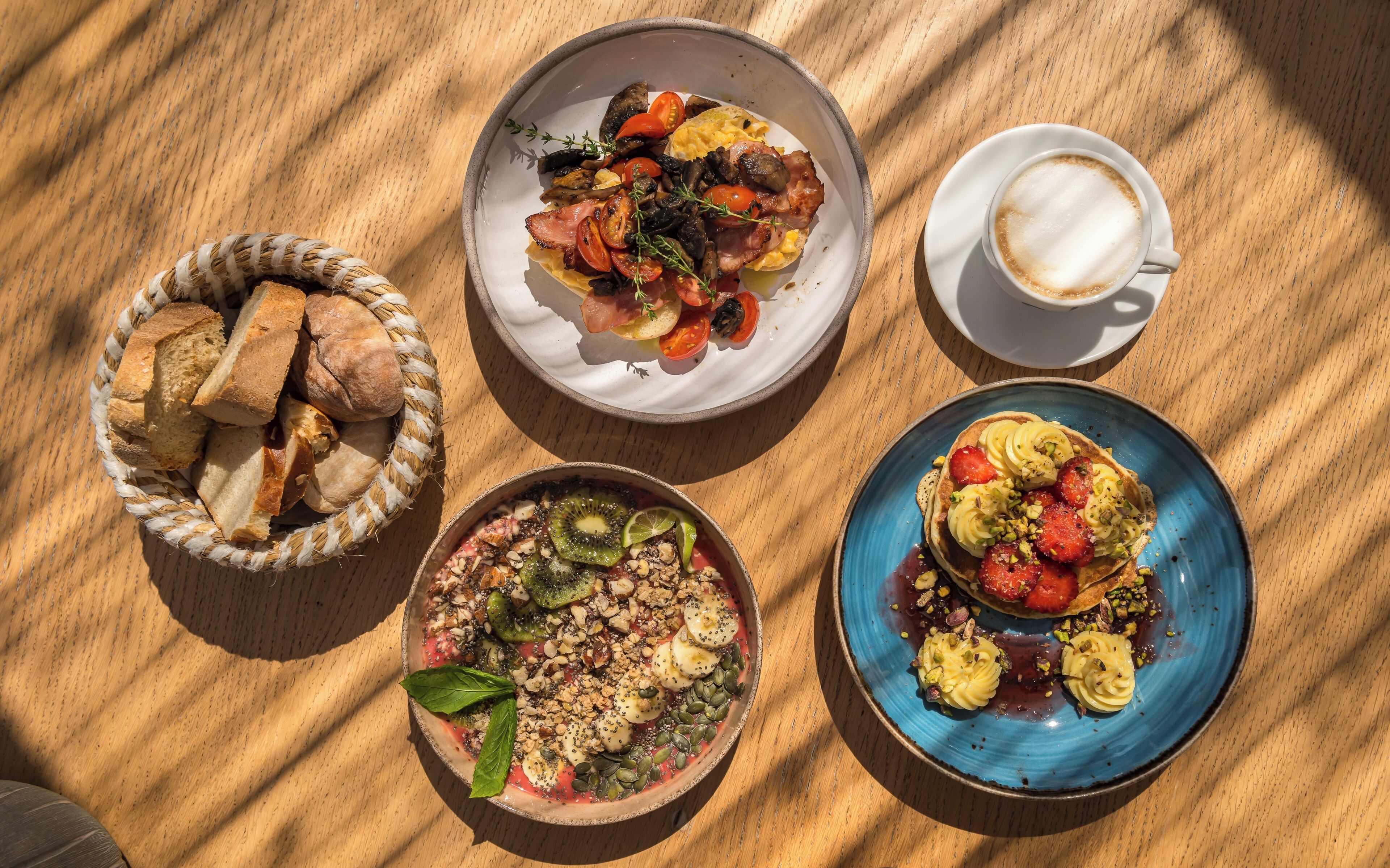 Een tafel met vier gerechten: een broodmand, een salade met tomaten en champignons, een fruitsalade en een stapel pannenkoeken met aardbeien en noten.