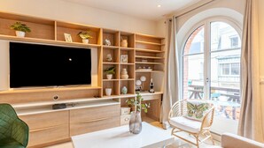 A modern living room with a large flat-screen TV, wooden shelving, a dining table, and a view of the outside balcony.