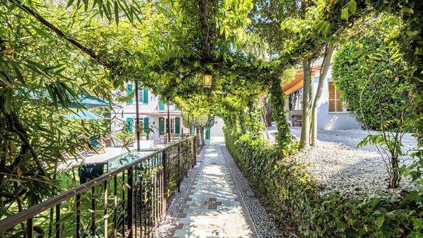 Un chemin bordé de verdure et muni d’une rambarde métallique, qui mène à un bâtiment à la façade blanche et aux volets verts.