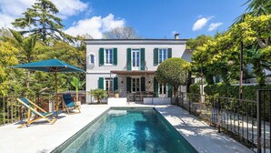 Une piscine rectangulaire avec une terrasse blanche, un bâtiment à deux étages doté de volets verts et un parasol vert.