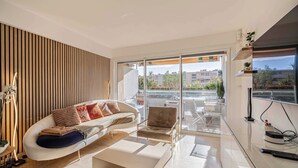 A modern living room with a sofa, armchair, and coffee table, large sliding glass doors leading to a balcony with plants and a city view.