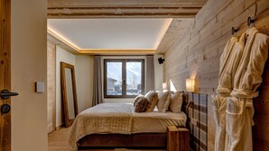 Une chambre avec un plafond en bois, un grand lit recouvert d’un tapis en fourrure, une table de chevet en bois et une fenêtre donnant sur des bâtiments enneigés.