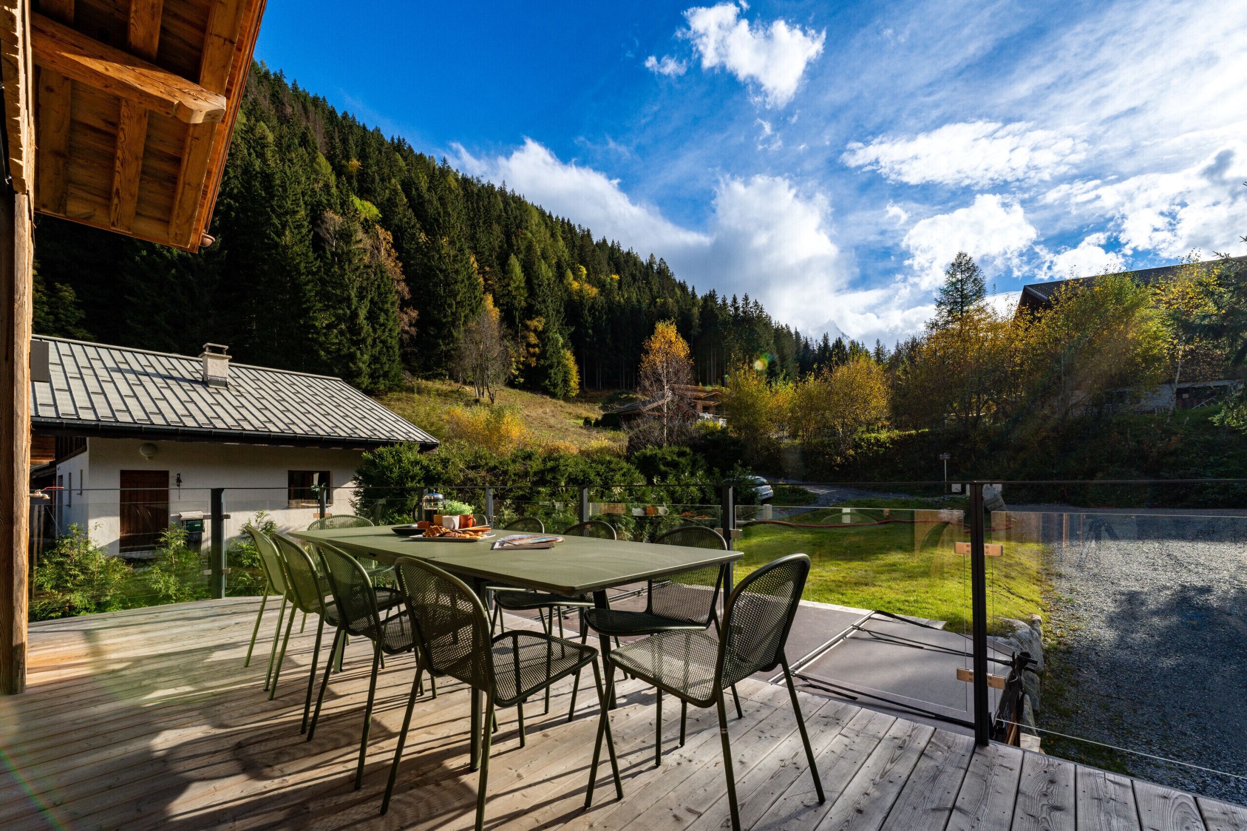 A wooden deck with a table and chairs, overlooking a forested hillside and a body of water.