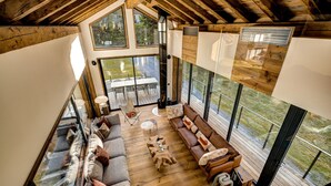 A spacious living area with a wooden floor, a brown leather sofa, a glass coffee table, and a large window offering a view of the outdoors. - Serviced Luxury Chalet with 10m Indoor Pool (Chamonix-Mont-Blanc)