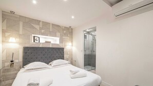 A modern hotel room with a large bed, two bedside lamps, a grey upholstered headboard, and a glass shower enclosure. - Beautiful apartment - Croisette Gray Albion (Cannes)