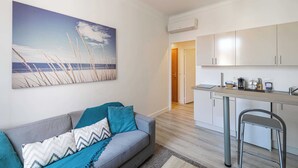 Ein modernes Wohnzimmer mit grauem Sofa, einem Strandbild und einer Küchenzeile mit Schränken und einem Barhocker.