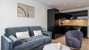 Uma sala de estar moderna com um sofá cinzento, uma poltrona roxa e uma mesa de centro branca. Há uma área de cozinha com armários pretos e uma bancada de madeira visível através de uma porta aberta.
