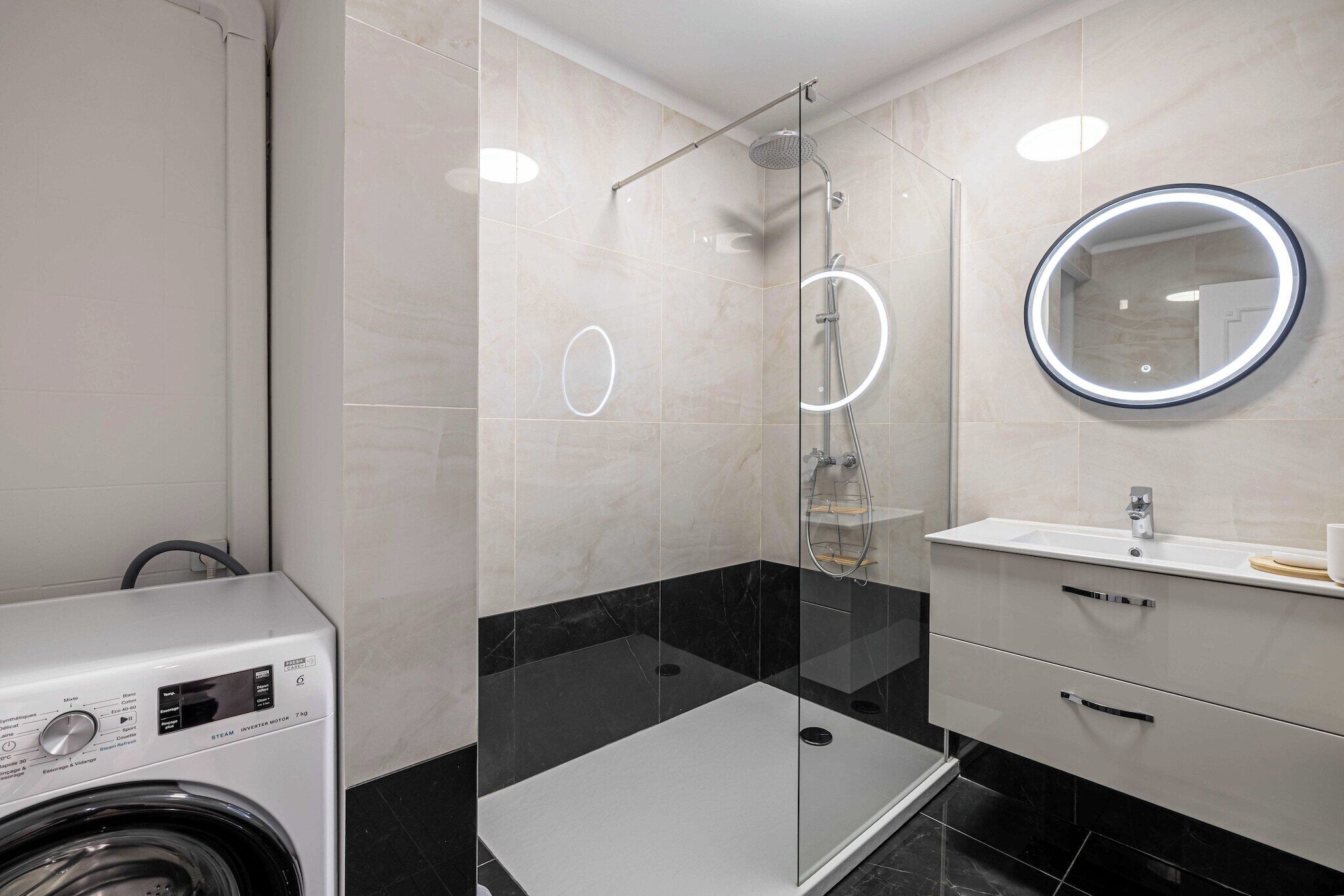 A modern bathroom with a glass-enclosed shower, a round mirror, a white sink, and a built-in washing machine.