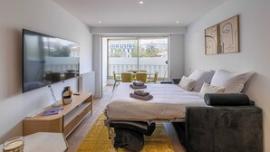 Quarto de hotel moderno com cama, mesa de cabeceira, televisão e varanda com cadeiras.