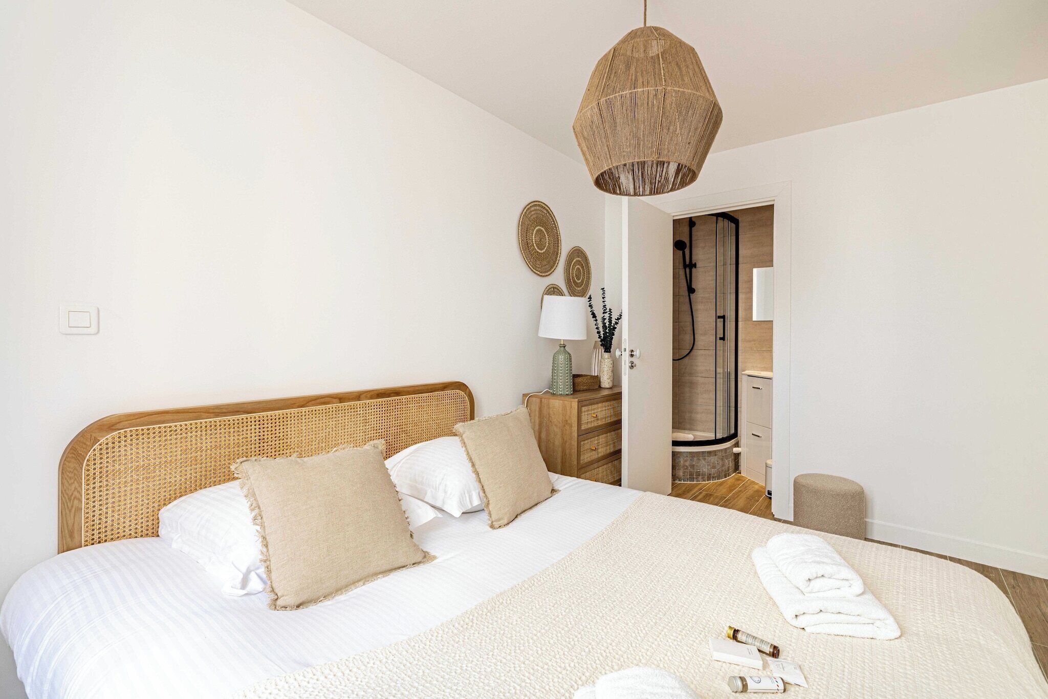 A bedroom with a large bed, a wooden headboard, a hanging light fixture, a bedside table with a lamp, and a bathroom visible through an open door.