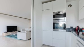 Una cocina moderna con electrodomésticos integrados, un comedor con mesa blanca y sofá, y un televisor de pantalla plana grande.