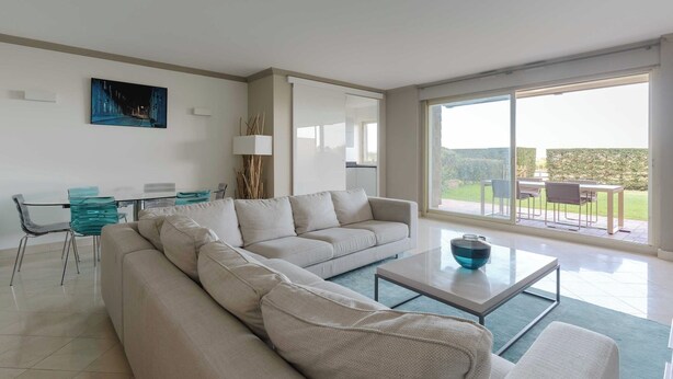Una sala moderna con un sofá grande, una mesa de centro y una zona de comedor con sillas y una mesa. Una ventana grande ofrece vistas al exterior.
