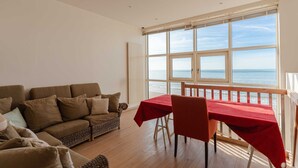 Un salon avec une grande fenêtre donnant sur la mer, une table rouge et un canapé marron.