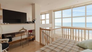 Une chambre avec une grande fenêtre donnant sur la mer, un lit, une télévision et un petit coin salon avec une table et des chaises.