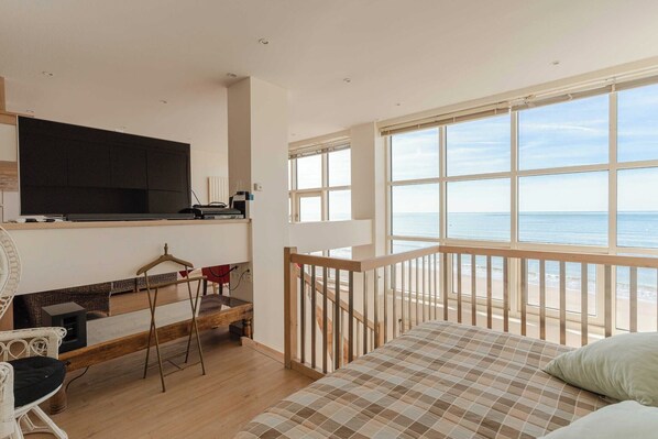 A bedroom with a large window overlooking the sea, a bed, a TV, and a small seating area with a table and chairs.